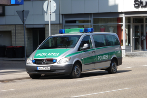 Ein Polizeifahrzeug parkt am Straßenrand vor einem Gebäude mit Glastüren, auf denen Schaufensterpuppen zu sehen sind. Im Hintergrund sind Schilder und Mülltonnen zu erkennen.