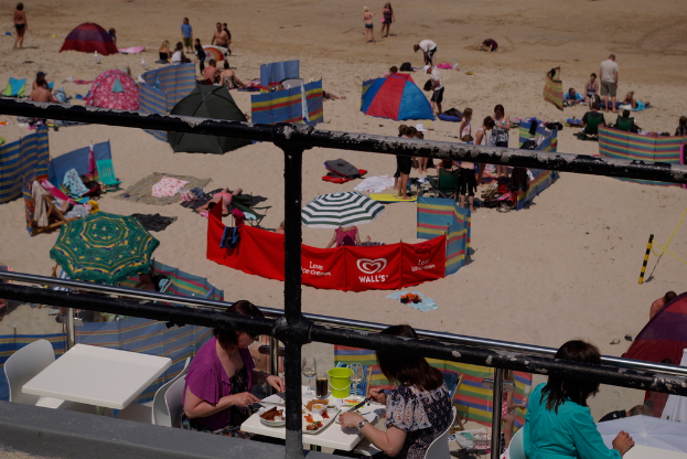 Eine schwarze Brüstung im Vordergrund mit einem leeren Tisch und Stühlen, zwei Frauen sitzen am Tisch und essen, Sand, ein rotes Tuch und einige Menschen stehen rechts im Hintergrund, mit Zelten und Kleidung weiter hinten.