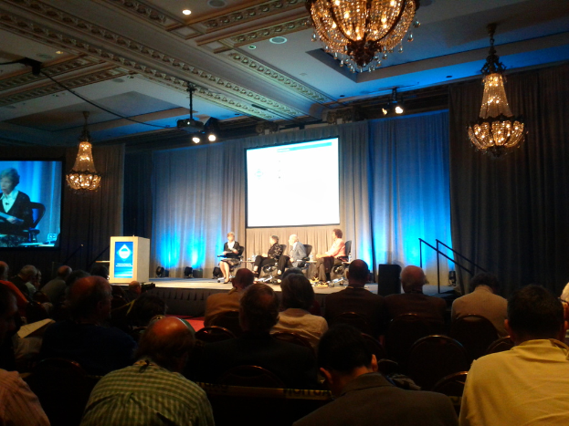 Eine Gruppe von Menschen auf Stühlen in einem Raum mit einer Bühne, einem Podium, Scheinwerfern, Bildschirmen, Vorhängen, Kronleuchtern, einem Redner und einem Dach.