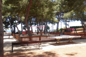 Eine Parkszene mit einem zentralen Tischtennistisch, umgeben von Bänken, einem Zaun, Schaukeln, Pfählen, Pflanzen und Bäumen, mit einem Gewässer und einem klaren blauen Himmel im Hintergrund.