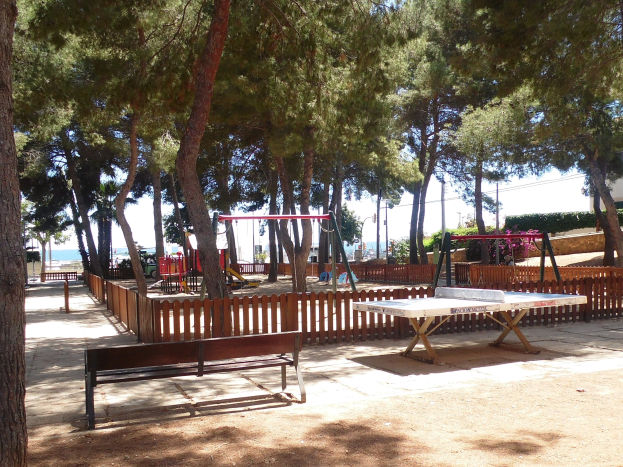 Eine Parkszene mit einem zentralen Tischtennistisch, umgeben von Bänken, einem Zaun, Schaukeln, Pfählen, Pflanzen und Bäumen, mit einem Gewässer und einem klaren blauen Himmel im Hintergrund.