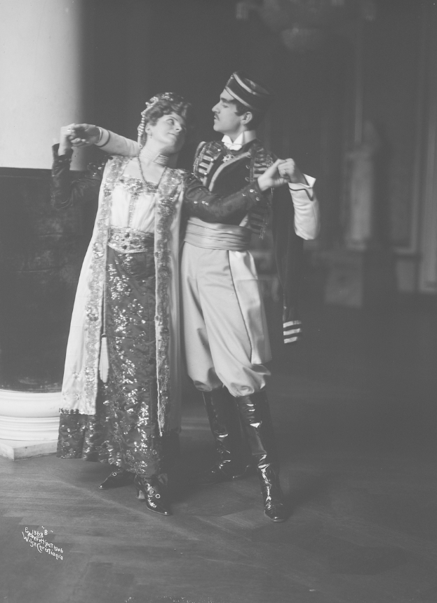 Ein Mann und eine Frau in Abendgarderobe tanzen in einem Ballsaal, mit einer Säule und einer Wand im Hintergrund, während einer Vorstellung.