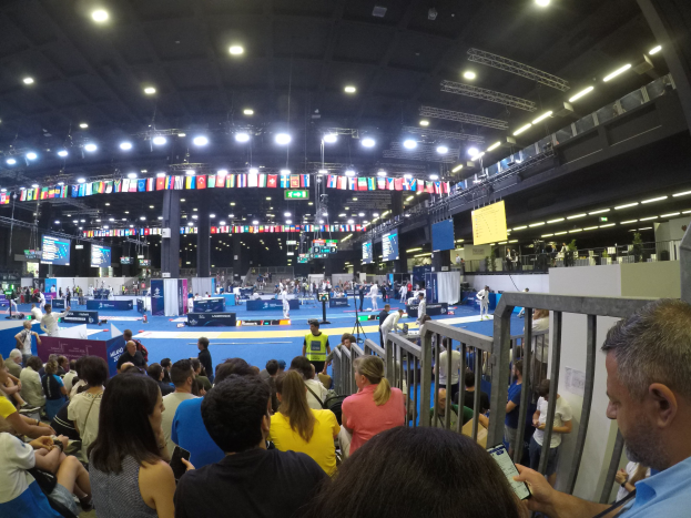 Eine große Menschenmenge sitzt und steht in einer Indoor-Sportarena, mit Bildschirmen, Fahnen und Deckenleuchten im Hintergrund.