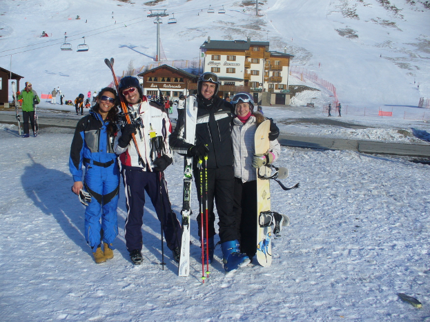 Vier Personen posieren für ein Foto mit Skaterausrüstung und Werkzeugen, mit Gebäuden und anderen Menschen im Hintergrund, vor einer schneebedeckten und vereisten Berglandschaft.