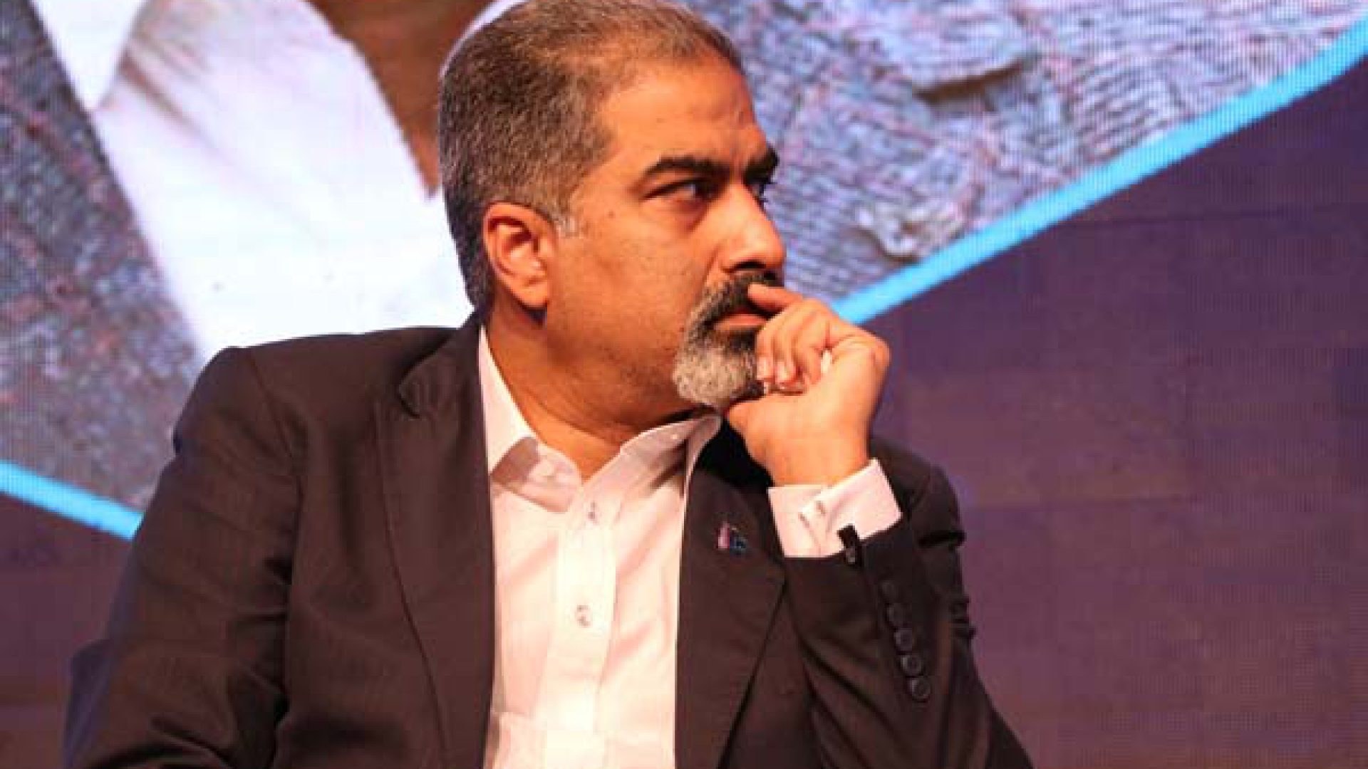 Ein Mann im Anzug sitzt auf einer Bühne, in Gedanken versunken, einem Bildschirm zugewandt.