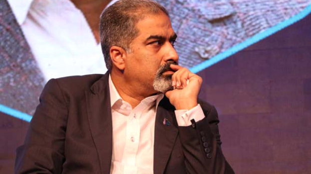 Ein Mann im Anzug sitzt auf einer Bühne, in Gedanken versunken, einem Bildschirm zugewandt.