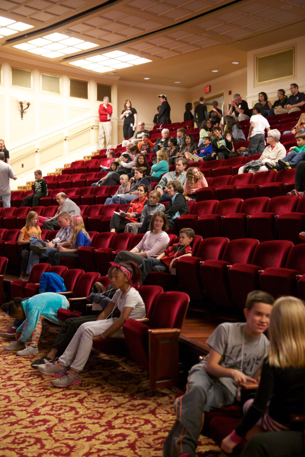 Eine große Gruppe von Menschen, darunter Kinder, die auf Stühlen sitzen und stehen, in einem Theater mit herumstehenden Stühlen und einer beleuchteten Wand im Hintergrund unter Deckenbeleuchtung.