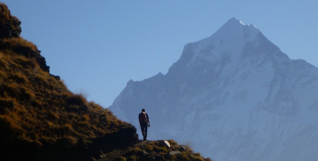Ein Mann mit einem Rucksack geht auf einem geneigten Pfad, mit einem Berg und Himmel im Hintergrund.