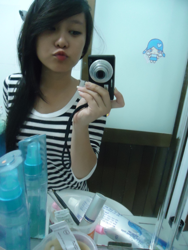 Eine Frau steht vor einem Schminktisch mit Kosmetikartikeln und hält mit einer Kamera ein Selfie, während sie in einen Spiegel schaut, mit einer Tür im Hintergrund.