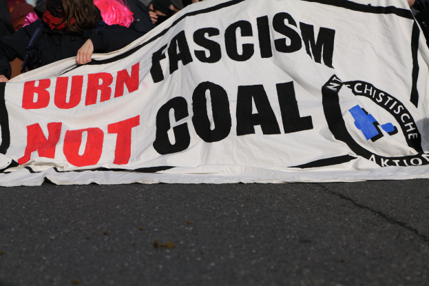 Eine Gruppe von Menschen steht auf einer Straße und hält ein buntes Banner mit der Aufschrift "Verbrenne Faschismus nicht Ziel."