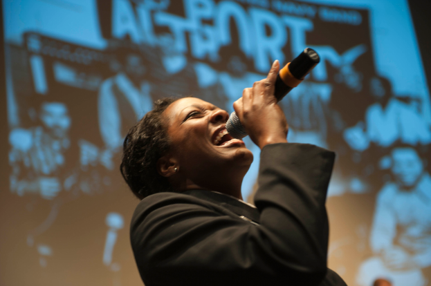 Eine Frau in einem schwarzen Kleid singt in ein Mikrofon auf der B├╝hne, ihre Augen sind geschlossen und ihr Gesicht von Scheinwerfern erhellt.