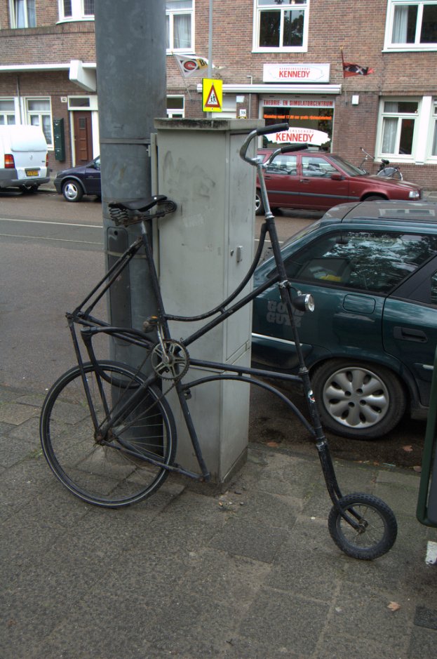 Ein Fahrrad steht in der Nähe einer Straße mit Pfählen, Namensschildern, Fahrzeugen, Gebäuden mit Fenstern und Vorhängen sowie Fahnen.