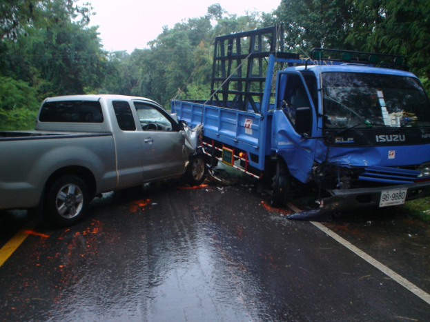Ein schwerbeschädigter Lastwagen mit eingedrückter Front und verbeulter Karosserie liegt auf der Seite einer Straße, umgeben von Bäumen unter einem klaren blauen Himmel.