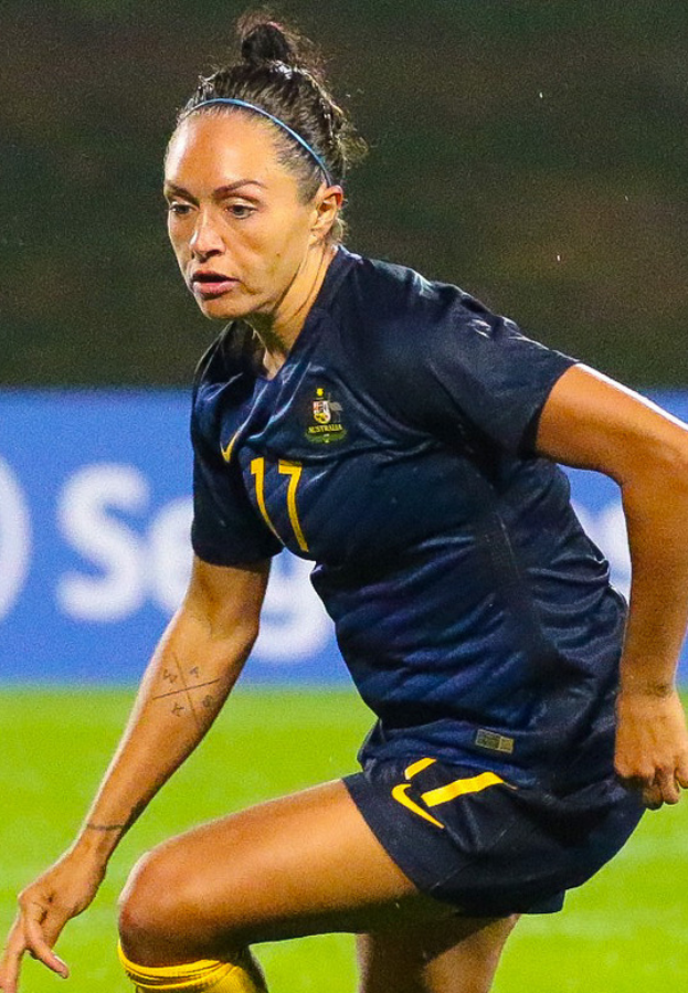 Eine Fußballspielerin in Aktion auf dem Feld, die ein blaues T-Shirt und gelbe Shorts trägt, mit einer blauen Tafel im Hintergrund.