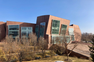 Großes mehrstöckiges Gebäude mit zahlreichen Fenstern, umgeben von Bäumen, Bänken, Gras und einem Laternenpfahl, mit anderen Gebäuden und einem klaren blauen Himmel im Hintergrund, identifiziert als der neue Campus der University of Illinois at Chicago.