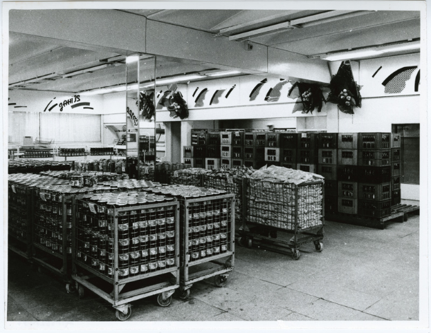 Ein Schwarz-Weiß-Foto eines Fabrikinnenraums mit fahrbereiten Lebensmitteln auf den Wagen auf dem Boden, Gegenständen auf den Regalen, Lampen an der Decke und Text an der Wand.