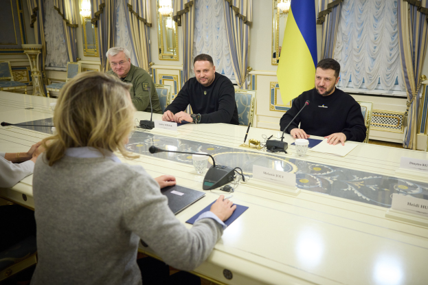 Eine Gruppe von Menschen, die um einen langen Tisch mit Mikrofonen, Namensschildern und anderen Gegenständen sitzen, in einer formellen Umgebung mit einer Flagge, Vorhängen, Spiegeln und Lichtern, die darauf hindeutet, dass sie Mitglieder des ukrainischen Parlaments sind, die sich zu einer Besprechung versammelt haben.