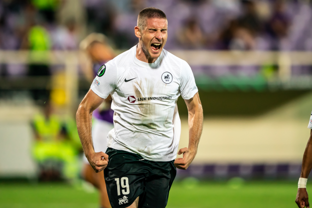 Ein Mann in weißem Hemd und schwarzen Shorts rennt auf einem Fußballfeld, feiert ein Tor und ist von einer Gruppe Menschen umgeben.