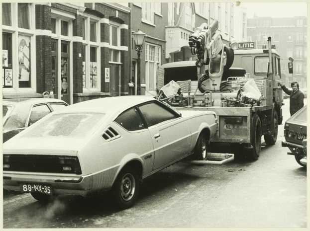 Ein Schwarz-Weiß-Foto eines Wagens, der von einem Abschleppwagen auf einer Straße mit anderen Fahrzeugen, einer Person in der Nähe und Gebäuden mit Fenstern im Hintergrund abgeschleppt wird.