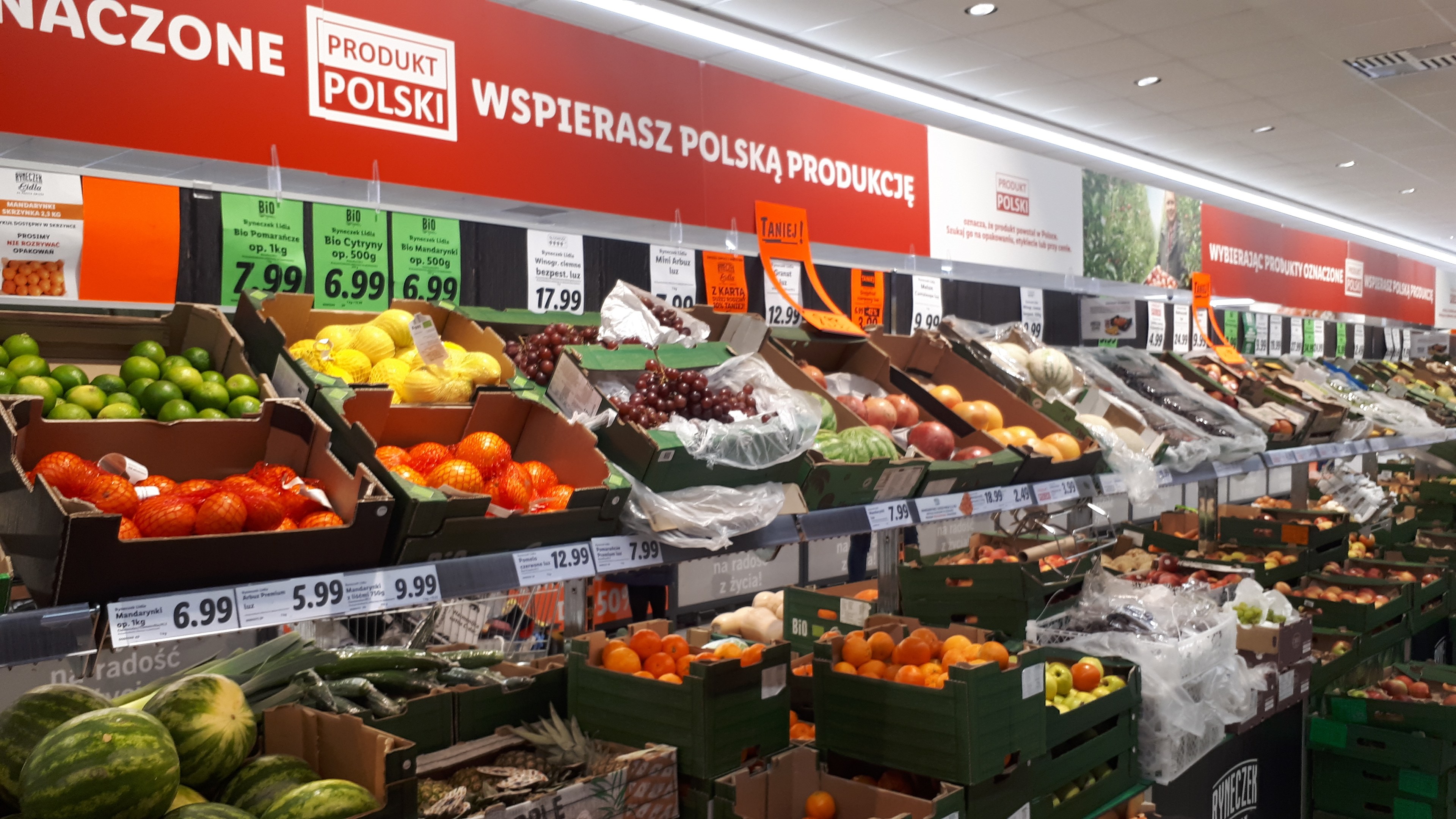 Ein Geschäft mit verschiedenen Obst- und Gemüsesorten in Kartons mit Preisschildern, Informationsschilder an den Wänden und Deckenbeleuchtung.