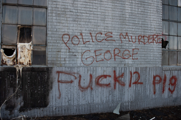 Ein Gebäude mit einer Wand, die mit der Aufschrift "Polizei Mord George Fuck 12 P19" beschmiert ist, mit Fenstern und verschiedenen Gegenständen davor.