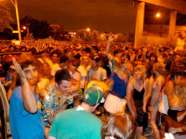 Menschen tanzen vorne bei einem Konzert, mit Lichtern, Pfählen, Bäumen und anderen Objekten im Hintergrund.