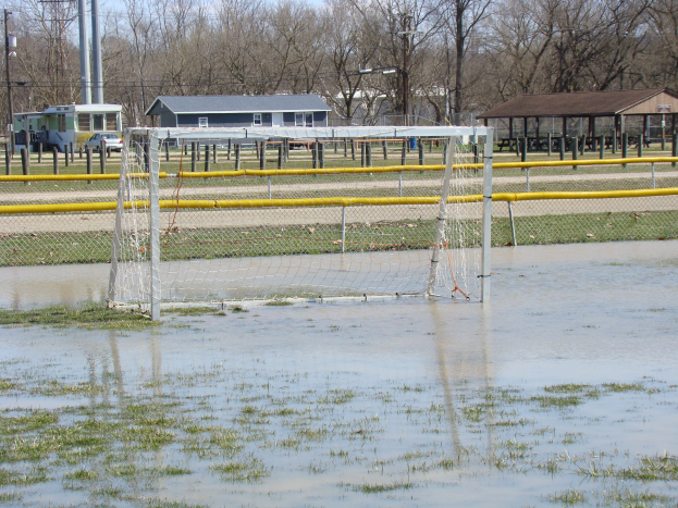 Ein Fußballtor steht in einem überfluteten Feld mit umliegender Wiese und Wasser, mit Hütten, Pfählen, Bäumen, Fahrzeugen und einem klaren blauen Himmel im Hintergrund.