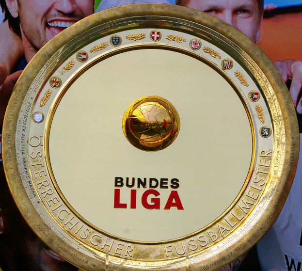 Ein Mann hält einen goldenen Schild mit der Aufschrift 'Bundesliga' hoch, im Hintergrund eine Fahne mit drei Personen.
