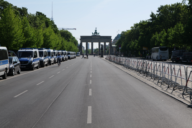 Eine Reihe von Polizeiwagen, die auf der Seite einer Straße vor dem Brandenburgertor in Berlin, Deutschland, geparkt sind, mit Menschen, die Fahrräder fahren und auf der Straße stehen, Barrieren, Bäumen und einem Bogen mit Statuen im Hintergrund.