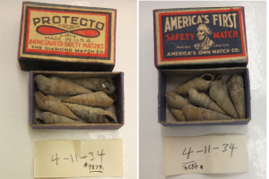 Zwei Streichholzschachteln mit der Aufschrift "Protecto" und "Amerikas erste Sicherheitszündholz" auf einem Tisch mit verschiedenen Inhalten und zwei nummerierten Papieren darunter.