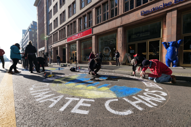 Menschen sitzen vor einem Gebäude mit Fenstern und Namensschildern auf dem Boden, umgeben von Flaschen und anderen Gegenständen, während sie an einer Klimademonstration in Berlin teilnehmen, die von Bäumen und einem klaren blauen Himmel umgeben ist.
