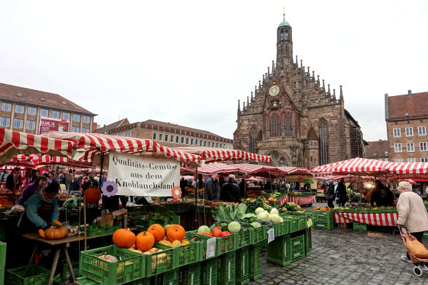 Ein belebter Markt in Nürnberg, Deutschland, mit verschiedenen Obst- und Gemüsesorten, Menschen beim Einkaufen, Zelten, Gebäuden mit Fenstern und einem Uhrenturm im Hintergrund unter einem sichtbaren Himmel.