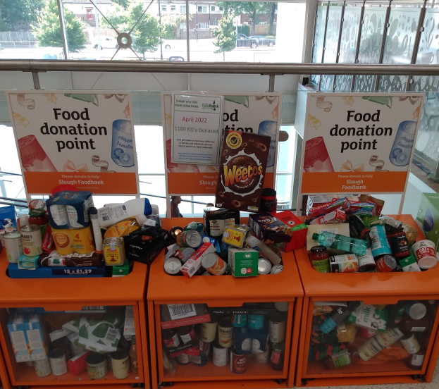 Orange Schübe mit Lebensmittelspenden gefüllt, darunter Flaschen, Dosen und Kisten, mit Informationsschildern und Blick auf Fahrzeuge, Bäume, Gebäude und Himmel durch Glasfenster.