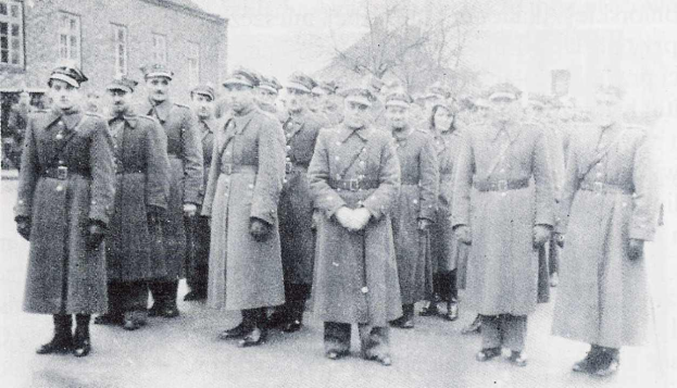 Eine Gruppe von Männern in deutschen Weltkriegs-Militäruniformen steht in einer Reihe vor einem Gebäude mit Fenstern, Bäumen und einem klaren Himmel im Hintergrund, dargestellt in Schwarz-Weiß.