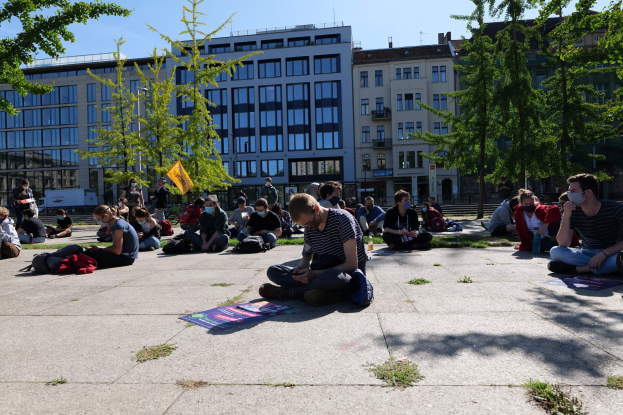 Eine Gruppe von Menschen sitzt vor einem Gebäude auf dem Boden während einer Demonstration in Berlin, einige tragen Masken, mit verstreuten Taschen und Gegenständen, unter einem klaren blauen Himmel umgeben von Bäumen.
