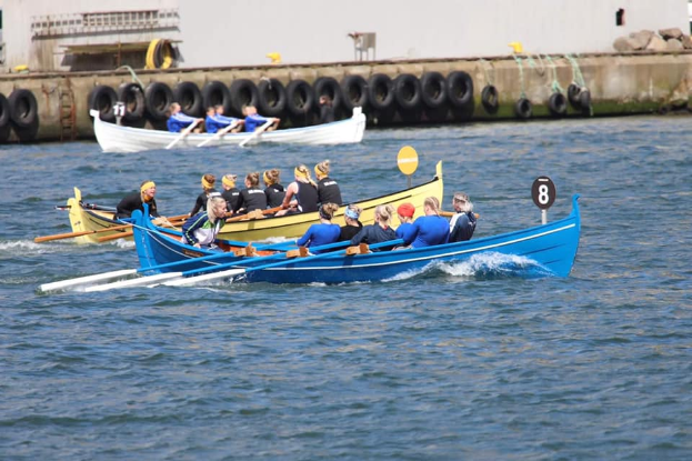 Eine Gruppe von Menschen in einem blauen und gelben Boot auf dem Wasser, die Paddel halten, mit einer Wand aus Reifen und einem Gebäude im Hintergrund.