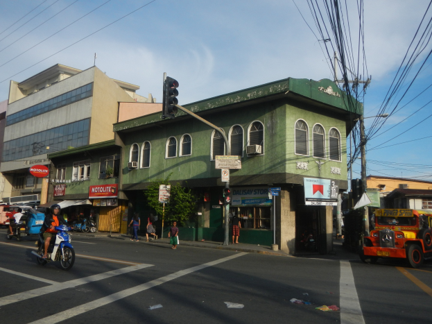 Eine belebte Stadtstraße mit einem grünen Gebäude im Hintergrund, fahrende Fahrzeuge, Fußgänger, eine Ampel an einem Mast, bewölkter Himmel und Stromleitungen auf Masten.