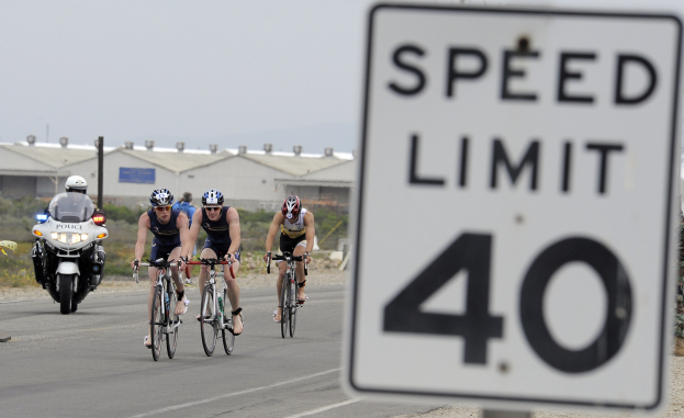 Eine Gruppe von Radfahrern mit Helmen fährt auf einer Straße neben einem Geschwindigkeitsbegrenzungsschild vorbei, mit Gebäuden, Masten, Pflanzen, Bäumen und einem klaren blauen Himmel im Hintergrund.