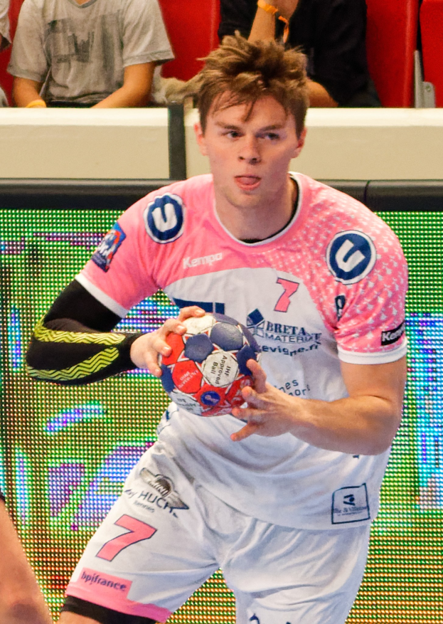 Ein Mann in einem pinken Hemd mit einem Fußball, daneben ist die Hand einer anderen Person zu sehen, während im Hintergrund Menschen auf Stühlen sitzen.