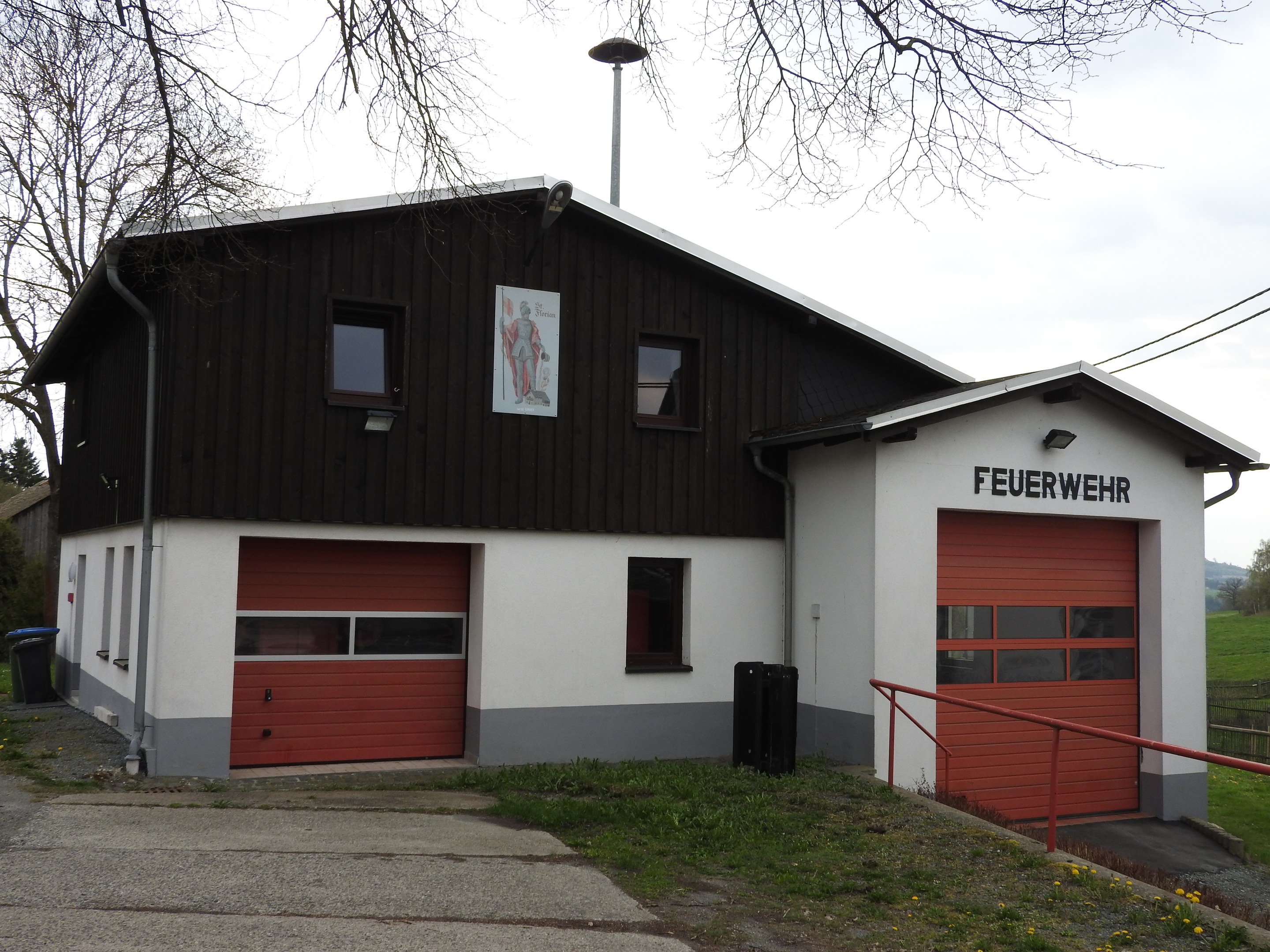 Rote und weiße Feuerwache mit Straße, Gras, gelben Blumen und Bäumen vor einem Himmel.