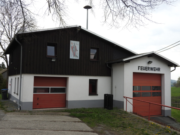Rote und weiße Feuerwache mit Straße, Gras, gelben Blumen und Bäumen vor einem Himmel.