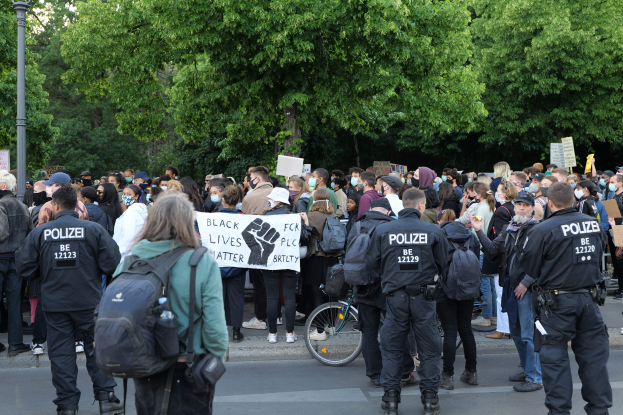 Eine große Gruppe von Menschen nimmt an einer Black-Lives-Matter-Demonstration auf einer Straße in Berlin teil, einige halten Schilder und andere tragen Mützen und Taschen, mit einem Fahrrad im Vordergrund und Bäumen und einem Pfahl im Hintergrund.