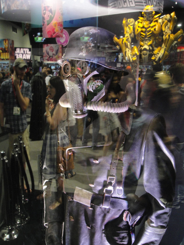 Ein reflektierender Spiegel in einer Halle, der eine Person in Anzug und Helm in der Vordergrund zeigt, eine Gruppe von Menschen, Stangen, Plakate mit Text, Bilder, eine gelbe Spielzeugfigur und verschiedene andere Gegenstände.