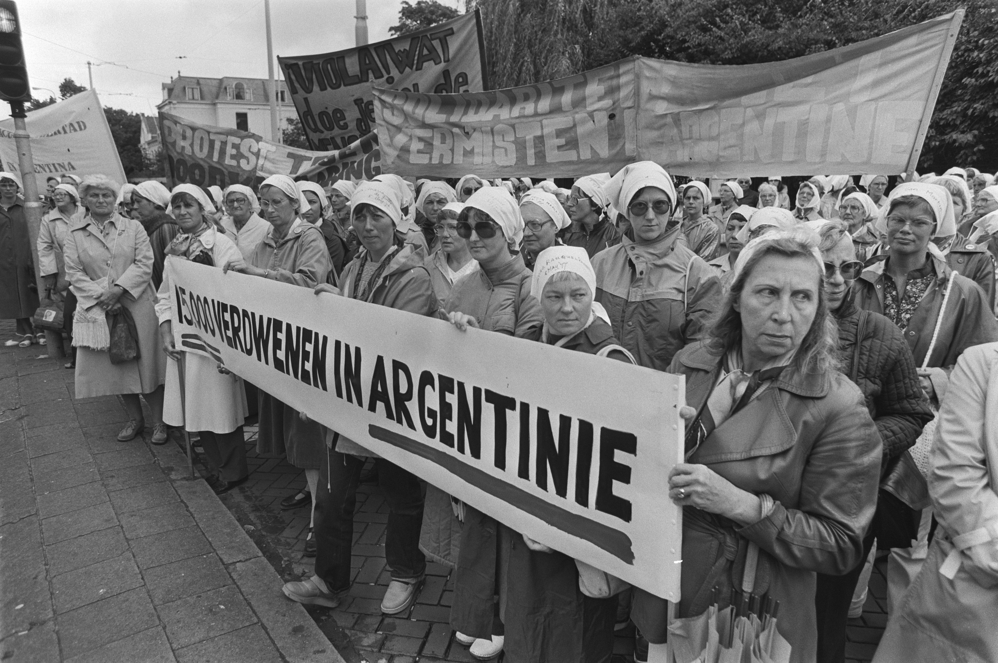 Eine Gruppe von Frauen hält ein Banner mit der Aufschrift "Argentinischer Frauenmarsch in Argentinien" vor einem Verkehrssignal, Bäumen, Gebäuden und einem bewölkten Himmel.