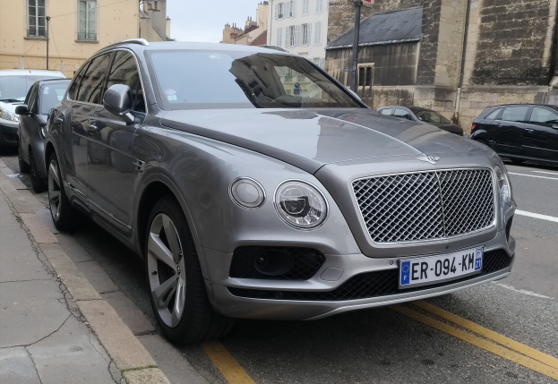Ein Bentley Bentayga V8 S fährt auf einer Straße mit einem Gehweg auf der linken Seite und Gebäuden mit Fenstern im Hintergrund, unter einem sichtbaren Himmel.
