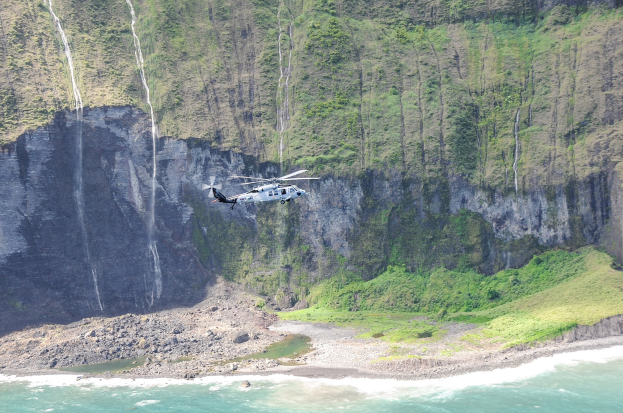 Ein Hubschrauber fliegt über das Meer in der Nähe einer Klippe, mit Wasser darunter, Felsen und Gras am Boden und Bäumen auf dem Berg im Hintergrund.
