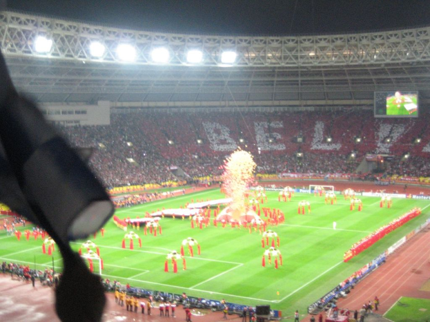 Ein Stadion mit Flutlicht, Eisenstäben, Zuschauern, stehenden Personen, Werbetafeln, Anzeigetafeln, Sicherheitskameras, Fahnen und Dekorationen.