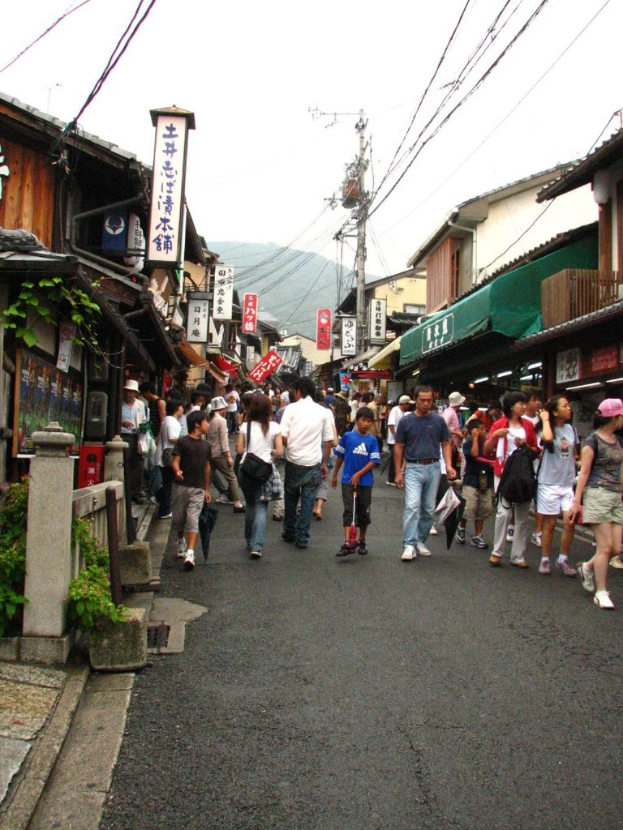 Eine Gruppe von Menschen, einige stehen und einige gehen auf einer Straße mit Plakaten, Häusern, Pflanzen, Pfählen und Kabeln in der Nähe.