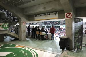 Eine Gruppe von Menschen steht vor einem sitzenden Publikum in einem Stadion während eines Boston Celtics vs New York Jets Ereignisses, mit Schildern, einem Müllcontainer, einer Tafel, einer Treppe mit Geländern und Deckenleuchten.
