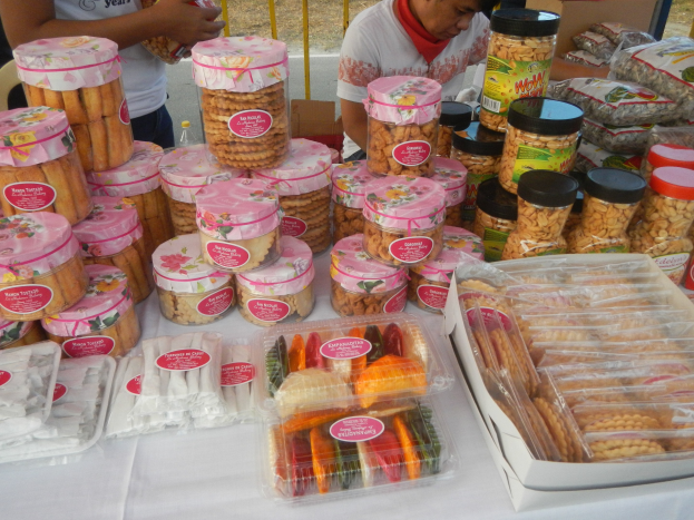 Eine Gruppe von Menschen um einen Tisch mit verschiedenen Lebensmitteln auf einem Food-Festival, mit einem Geländer und einer Straße im Hintergrund.